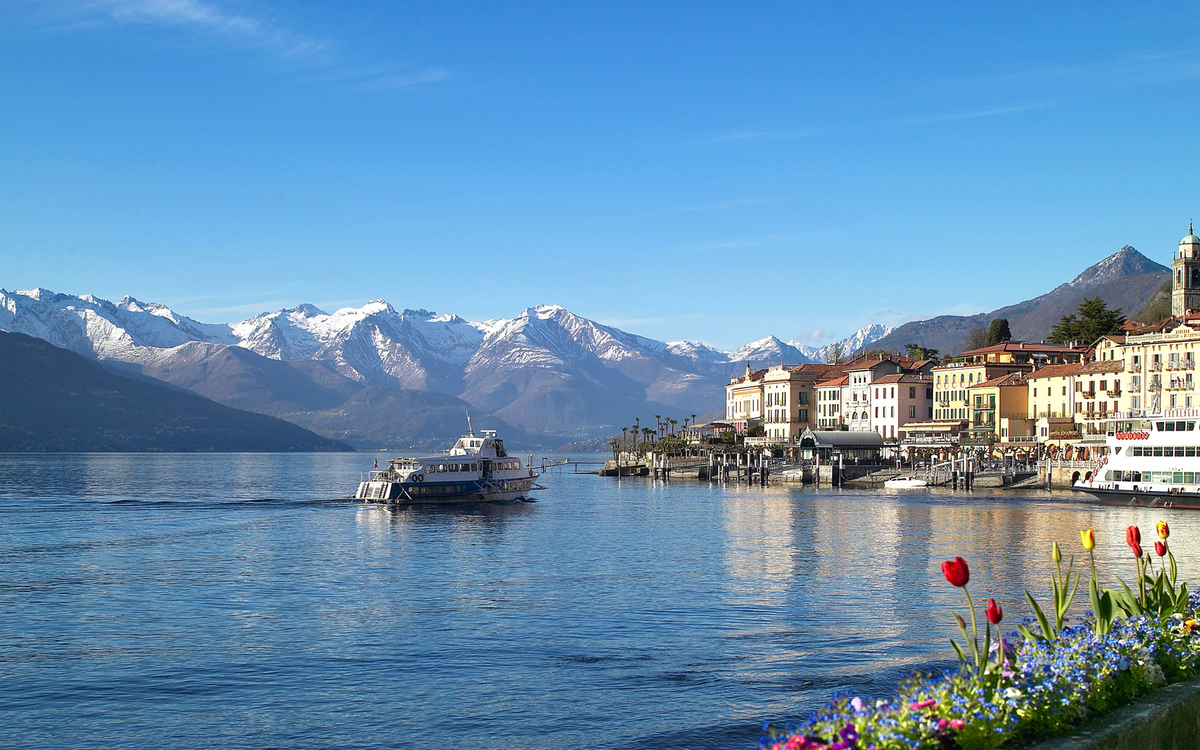 Stadt am See mit Bergen im Hintergrund und Blumen im Vordergrund.