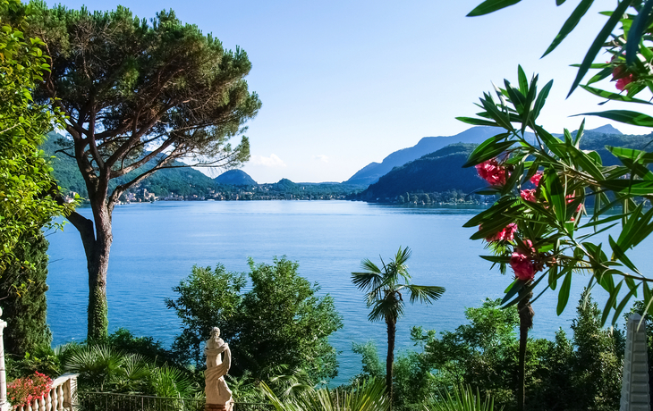 Blick auf einen See mit Palmen, Bergen und Statue im Garten.