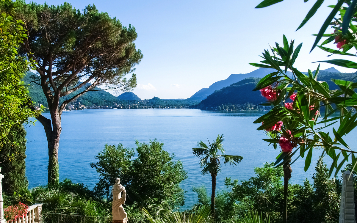 Blick auf einen See mit Palmen, Bergen und Statue im Garten.