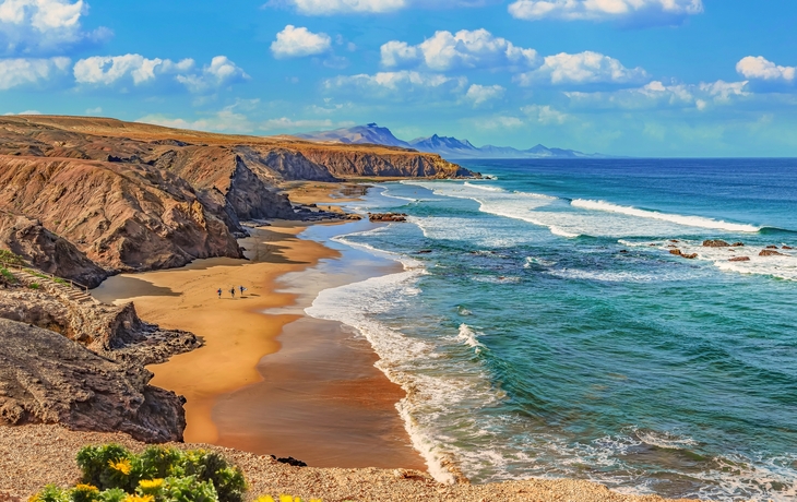 Küste mit Sandstrand, Felsen und blauem Meer unter wolkigem Himmel.