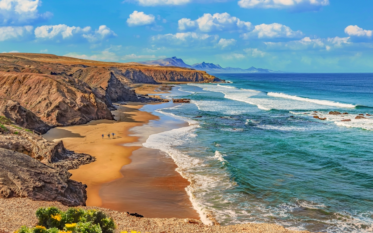 Küste mit Sandstrand, Felsen und blauem Meer unter wolkigem Himmel.