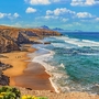 Küste mit Sandstrand, Felsen und blauem Meer unter wolkigem Himmel.