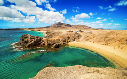 Küste mit blauem Meer und Sandstrand, Hügel im Hintergrund.