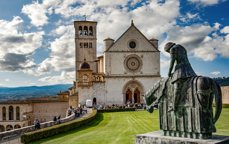 Kirche mit Glockenturm und Reiterstatue im Vordergrund bei blauem Himmel.