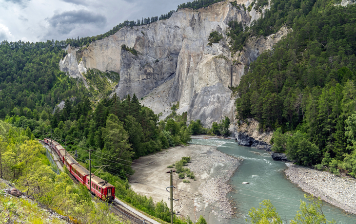 Roter Zug fährt durch eine bewaldete Berglandschaft entlang eines Flusses.