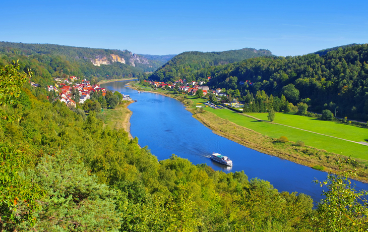 Fluss mit Boot, umgeben von bewaldeten Hügeln und Dorf im Hintergrund.