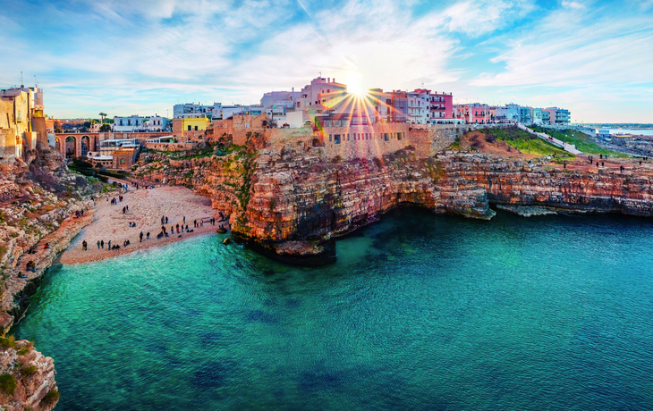 Küstenlandschaft mit Felsen, klarem Wasser und Gebäuden im Sonnenlicht.