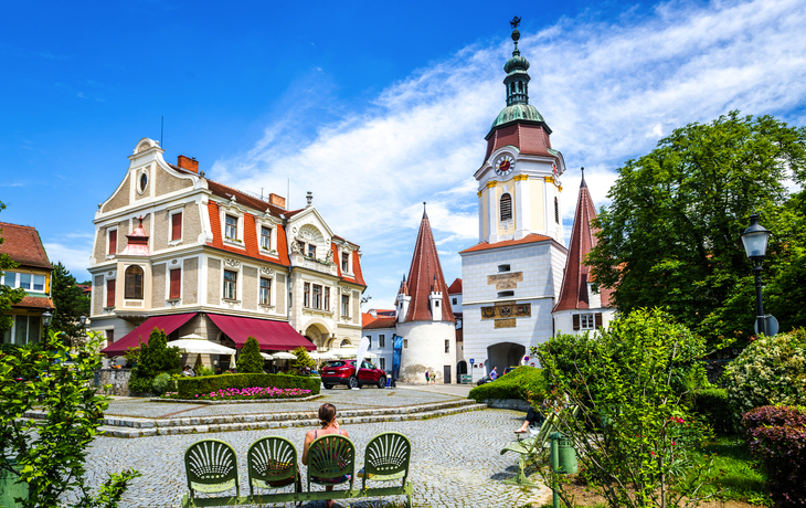 Historischer Platz mit Kopfsteinpflaster in einer malerischen Stadt, umgeben von traditionellen Gebäuden mit roten Dächern. Im Hintergrund ist eine Kirche mit einem Turm und Uhr sichtbar. Vorne sitzt eine Person auf einer Bank im Gartenbereich, der mit grünen Büschen und bunten Blumen dekoriert ist. Der Himmel ist klar und blau.