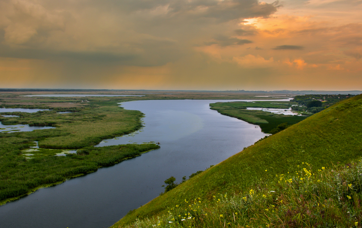 Flusslandschaft im Sonnenuntergang mit Wiesen und bewölktem Himmel