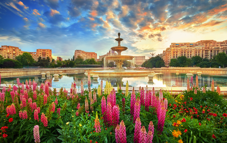Springbrunnen mit Blumenbeet im Vordergrund in einer städtischen Umgebung bei Sonnenuntergang.