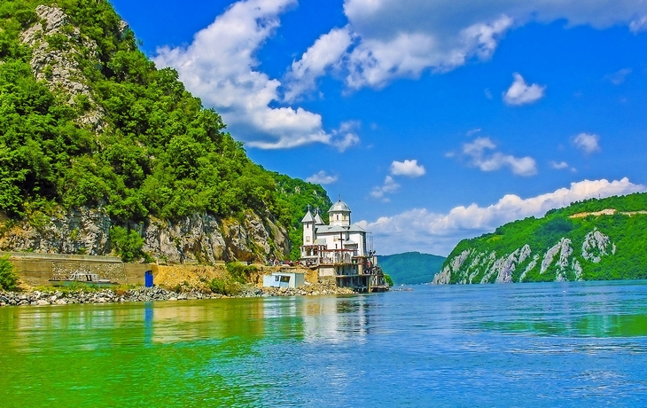 Flusslandschaft mit Kloster und bewaldeten Hügeln im Hintergrund.