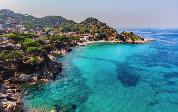 Küste mit blauem Meer und bewaldetem Hügel im Hintergrund.