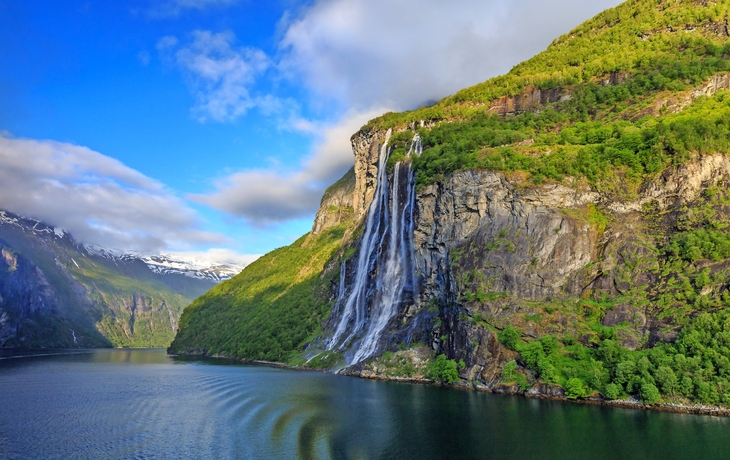 Wasserfall an steiler Felswand in einer grünen Landschaft unter blauem Himmel.