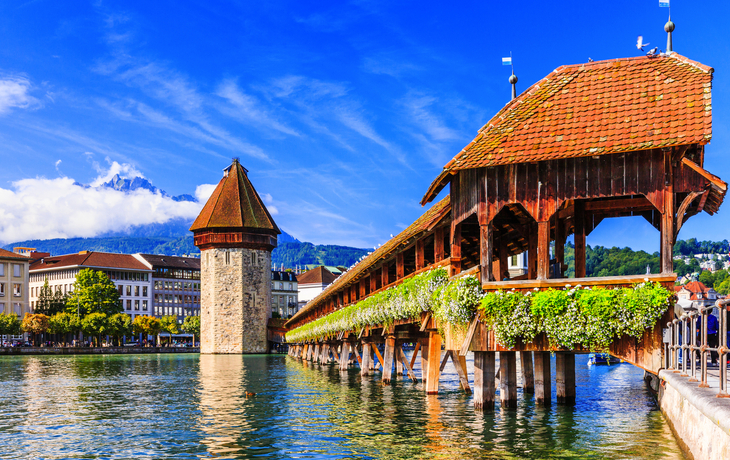 Holzbrücke mit Blumen und Wasserturm in Luzern vor blauem Himmel und Bergen im Hintergrund.
