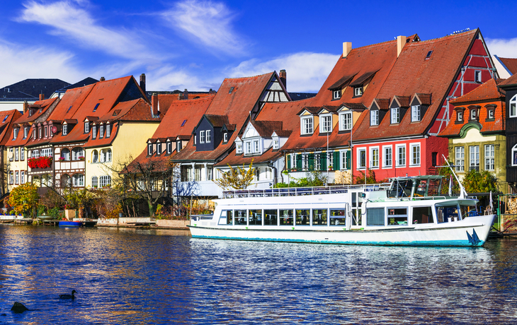 Bunte Fachwerkhäuser an einem Fluss mit einem weißen Touristenboot im Vordergrund.