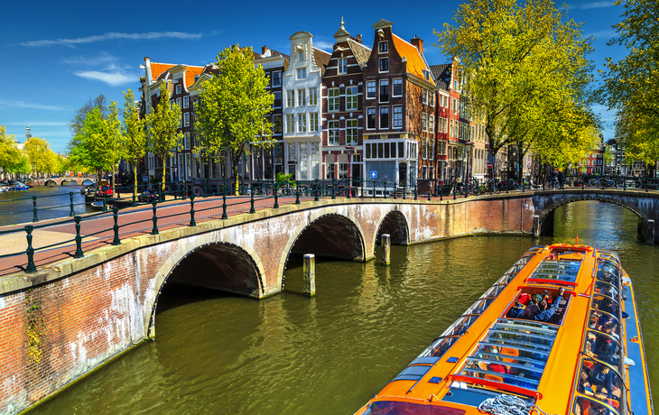 Kanal in Amsterdam mit Brücke, traditionellen Häusern und einem Touristenboot.