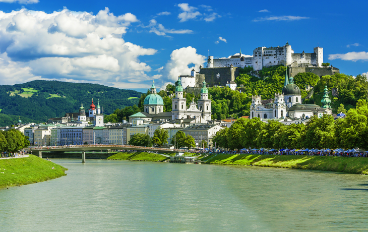 Flussblick auf Altstadt mit Festung Salzburg im Hintergrund, blauer Himmel mit Wolken.