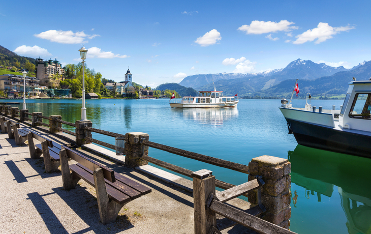Seeufer mit Booten und Dorf im Hintergrund, umgeben von Bergen.