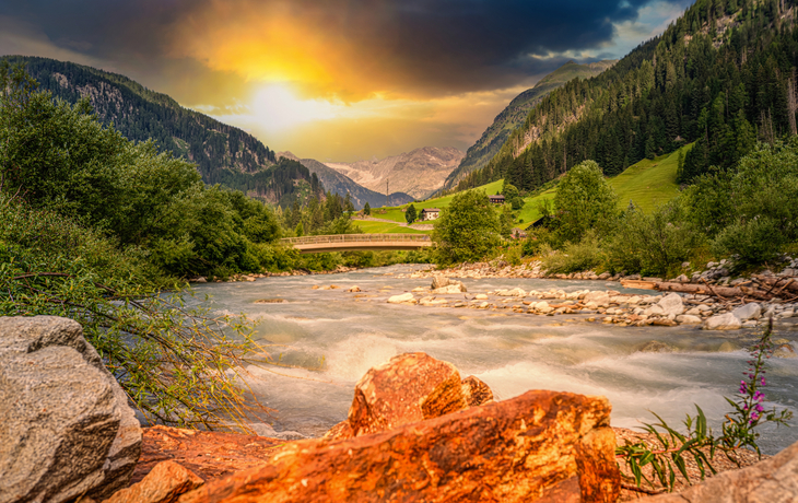 Fluss in Gebirgslandschaft bei Sonnenuntergang, umgeben von Bäumen und Bergen.