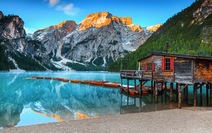 Holzhütte am klaren Bergsee mit Booten und Gebirge im Hintergrund bei Sonnenuntergang.