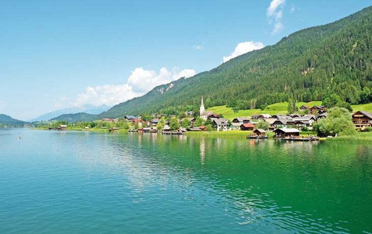 Dorf am Ufer eines Sees mit Bergen im Hintergrund an einem sonnigen Tag.