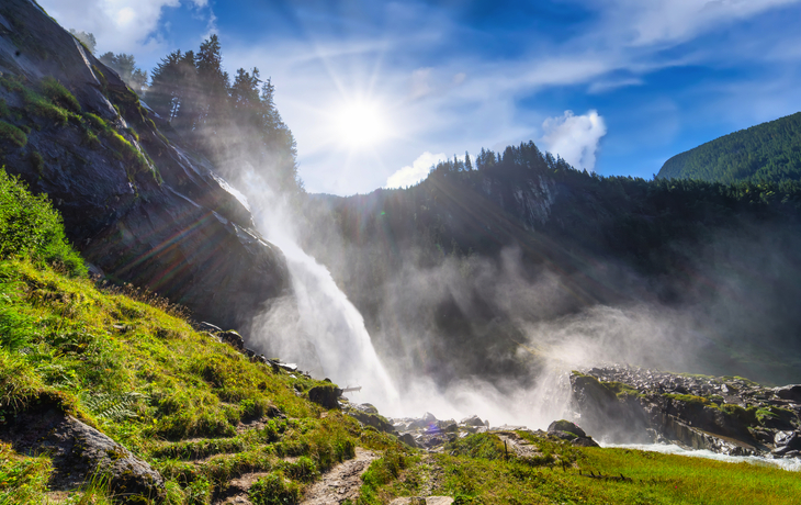 Sonnenbestrahlter Wasserfall in grüner Berglandschaft mit blauem Himmel.