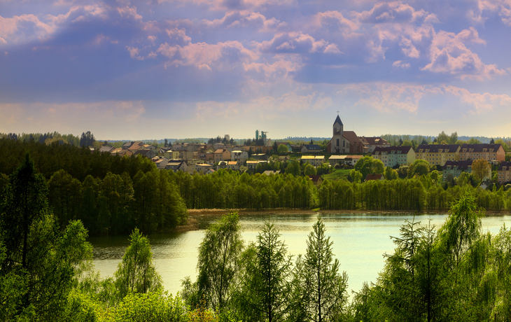 Stadtansicht mit Kirche und See im Vordergrund, umgeben von Wald und Wolken am Himmel.