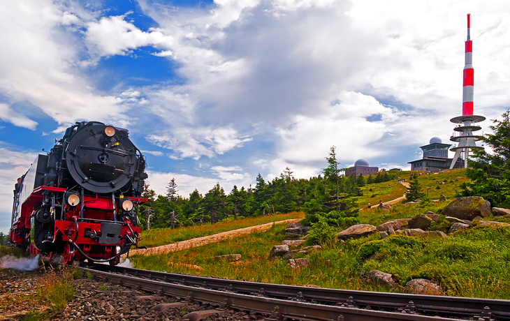 Dampflok fährt auf Schienen durch grüne Landschaft, im Hintergrund ein rot-weißer Sendeturm.
