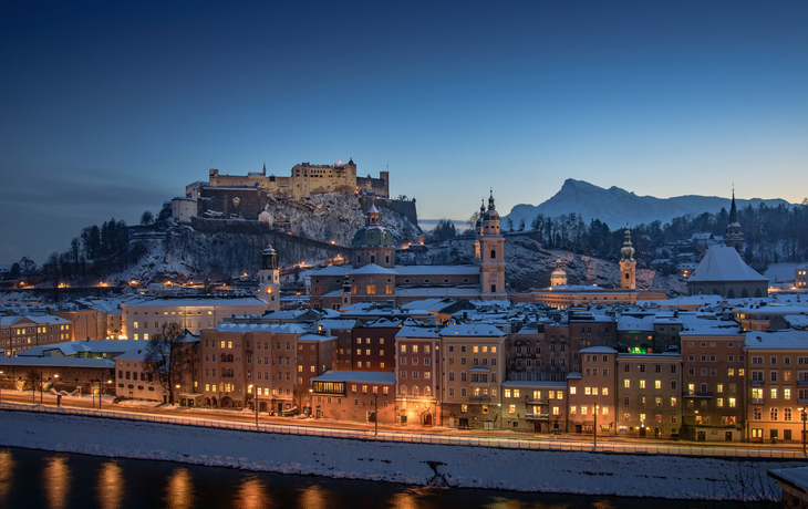 Verschneite Stadt und Burg bei Abenddämmerung, beleuchtet entlang eines Flusses.