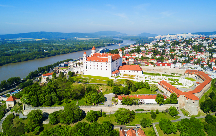Das Bild zeigt eine Luftaufnahme von Bratislava mit der Burg Bratislava, umgeben von grünen Parks, der Donau und einem Teil der Stadt im Hintergrund.