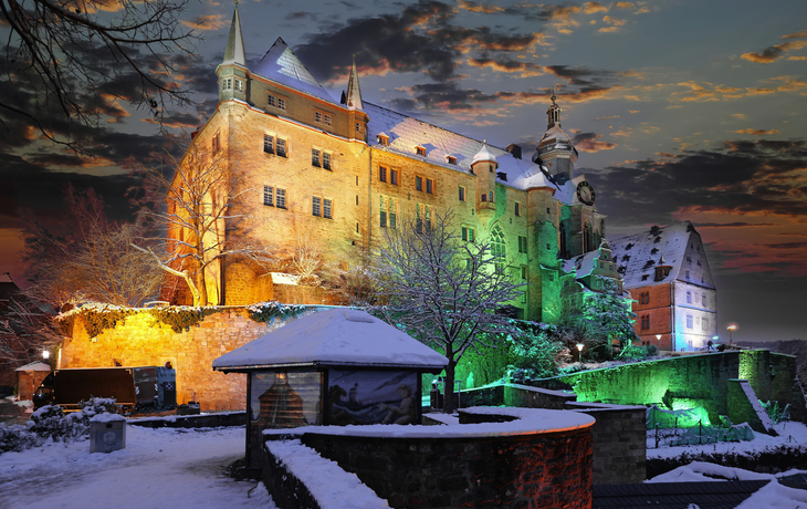 Beleuchtetes Schloss im Winter bei Sonnenuntergang, umgeben von Schnee und Bäumen.