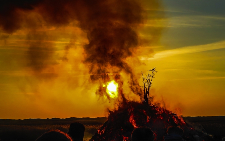 Menschenmenge beobachtet ein großes Lagerfeuer bei Sonnenuntergang, aus dem Rauch aufsteigt.