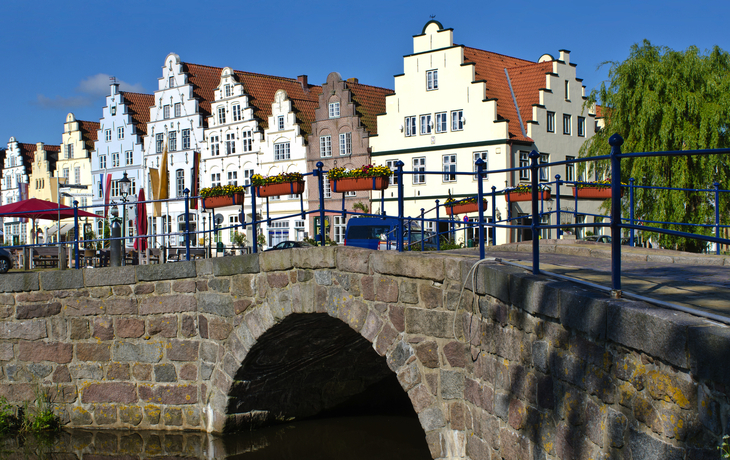 Brücke aus Stein vor historischen Giebelhäusern bei sonnigem Wetter.