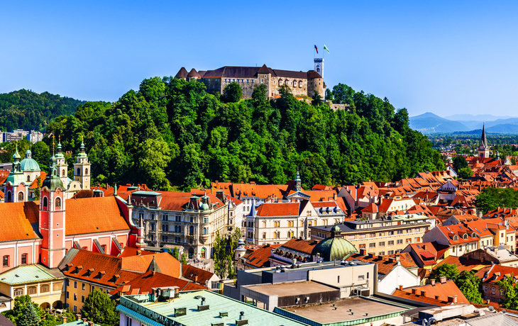 Altstadt von Ljubljana mit der mittelalterlichen Burg auf einem bewaldeten Hügel im Hintergrund.