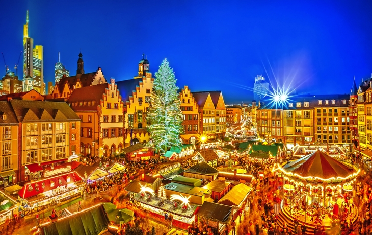 Belebter Weihnachtsmarkt mit Lichtern und geschmücktem Baum in einer Altstadt bei Nacht.