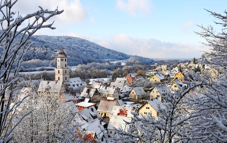 Verschneite Stadt mit Kirchturm und Hügeln im Hintergrund.