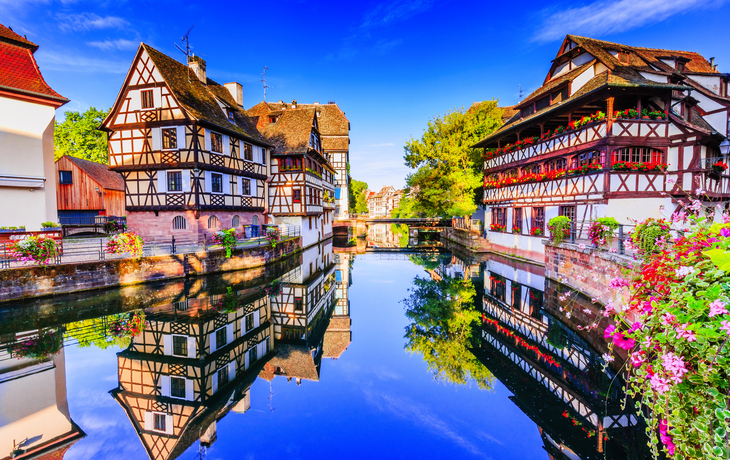 Fachwerkhäuser an einem Flussufer, spiegeln sich im Wasser, blauer Himmel.