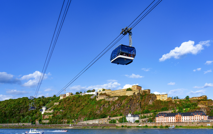 Seilbahn über einem Fluss mit Festung im Hintergrund bei blauem Himmel.