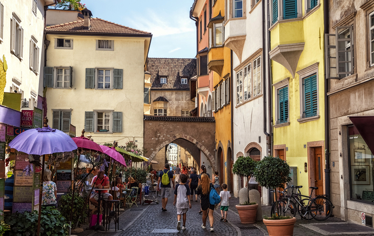 Menschen spazieren durch enge, bunte Altstadtstraße mit Straßencafé.