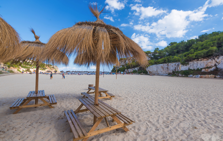 Holzpicknicktische mit Strohdächern an einem sonnigen Sandstrand.