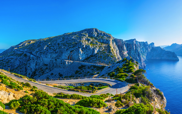 Küstenstraße mit scharfer Kurve, Berge und blaues Meer im Hintergrund.