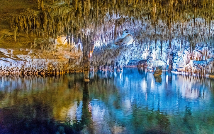 Eine farbenfrohe Höhle mit Stalaktiten und einem ruhigen Wasserbecken, das die Umgebung spiegelt.