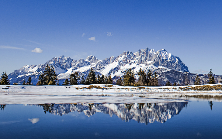 Verschneite Berge spiegeln sich in einem stillen See unter blauem Himmel.