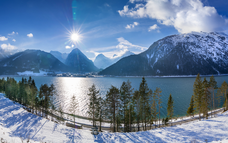 Verschneite Berglandschaft mit See und Sonne am Himmel.