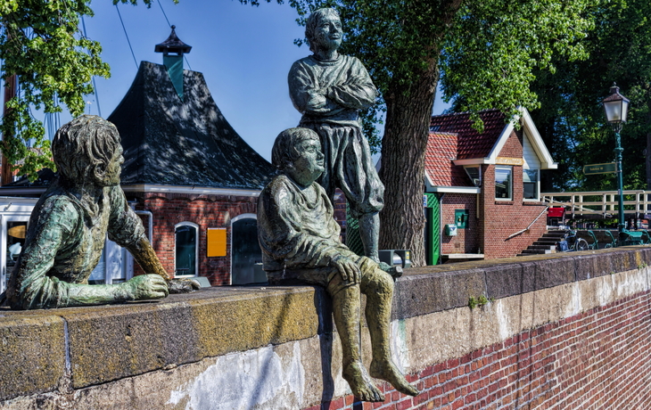 Bronzeskulpturen von Menschen auf einer Mauer vor Gebäude und Bäumen