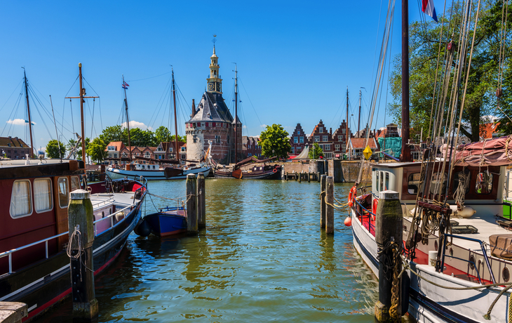 Historischer Hafen mit Schiffen und Turm im Hintergrund bei sonnigem Wetter.