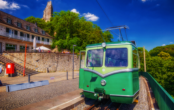 Grüne Bergbahn vor alter Ruine und Restaurant bei sonnigem Wetter.