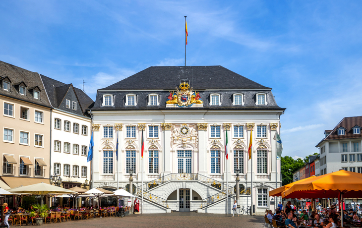 Historisches Gebäude mit Flaggen und Menschen auf einem Platz bei sonnigem Wetter.
