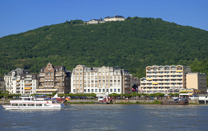 Gebäude am Flussufer mit einem bewaldeten Hügel im Hintergrund.
