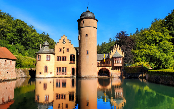 Historisches Wasserschloss mit Türmen und Bogenbrücke, umgeben von Wald.